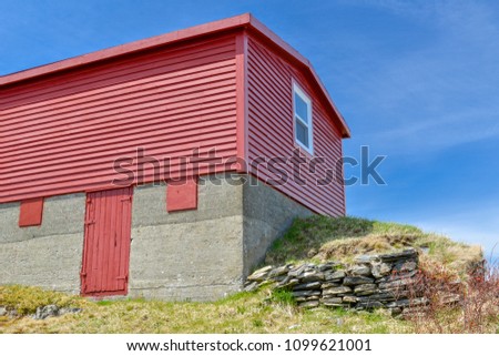 A red wooden storage shed with windows and a red wooden door. The sky in the background is bright blue with some light cloud covering. There's an old rock wall at the bottom of the picture. 
