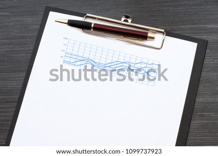 Black clipboard with white sheet of paper and metal pen on wooden table. Graphs and charts. Business concept