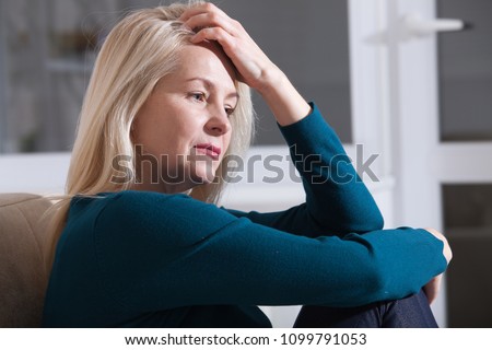 Sad depressed woman at home sitting on the couch, looking down and touching her forehead, loneliness and pain concept Royalty-Free Stock Photo #1099791053