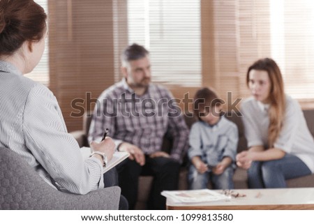 Rear view of female psychologist helping young family with a kid to solve child development problems. Family sitting on a sofa in the blurred background Royalty-Free Stock Photo #1099813580