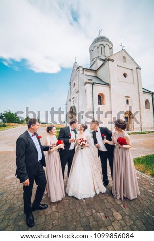 Cheerful and fun groom with bride, bridesmaids with groomsmen posing outdoors. Wedding moment of newlyweds. Old castle or chruch. Bridal day. People knocking glasses.