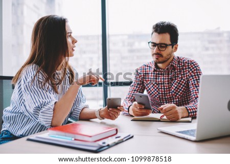 Male and female colleagues talking while share multimedia files via smartphones during working process at desktop, young employees discussing ideas while networking on mobile phones search information
