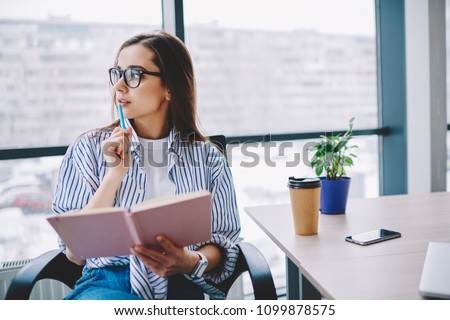 Pensive female author in eyewear looking away concentrated on idea for article sitting at desktop,skilled professional woman journalist pondering on publication holding pen and notebook in office Royalty-Free Stock Photo #1099878575