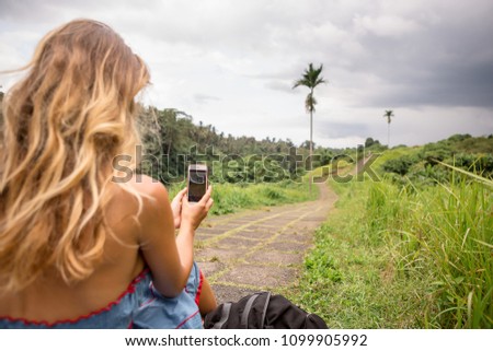 Beautiful young woman in blue dress seat at stone hill path in Asia and take pictures on her smartphone. Girl travel and explore world. Long lonely road in mountains. Travel and adventure mockup.