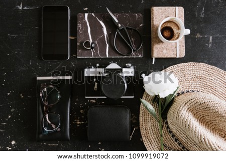 Travel essentials flat lay: vintage film camera, cork notepad, craft scissors, marble notepad, glasses, empty coffee cup, leather waller, hat, phone, peony, ring, black and white colors, table view