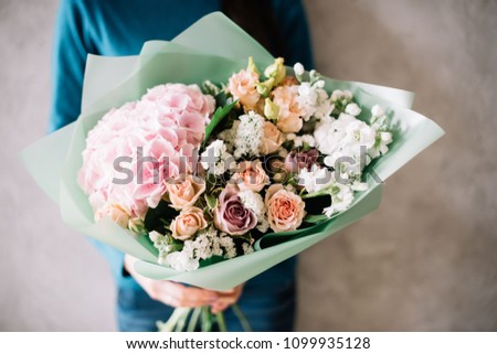 very nice young woman in a blue sweater holding a beautiful blossoming flower bouquet of fresh hydrangea, roses, carnations, matthiola, in pink and pastel cream colors on the grey wall background Royalty-Free Stock Photo #1099935128