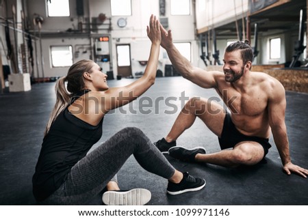 Two fit young people in sportswear smiling and high fiving together while sitting on a gym floor after working out Royalty-Free Stock Photo #1099971146
