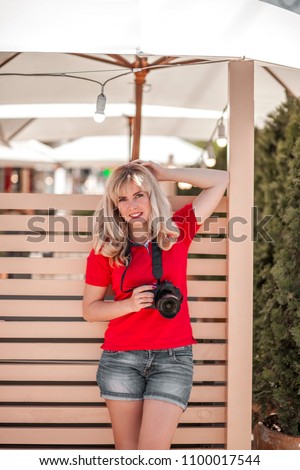 Portrait of beautiful smiling blonde girl, female, woman in red t-shirt, making photos at summer street. Portrait of professional photographer in casual wear holding photo camera.