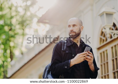 Outdoor view of thoughtful male tourist carries rucksack, holds modern smart phone in hands as tries to find right way, looks at online maps, focused somewhere away. People, travelling, technology