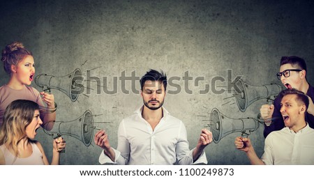 Impassive meditating young businessman paying no attention to crowd of screaming in megaphone angry people 