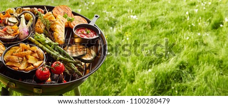 Assortment of fresh healthy vegetables on a BBQ grilling over a hot fire in a green grassy spring or summer field in banner format with copy space Royalty-Free Stock Photo #1100280479