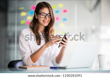 Young smiling business woman using smartphone near computer in office, copy space Royalty-Free Stock Photo #1100281091