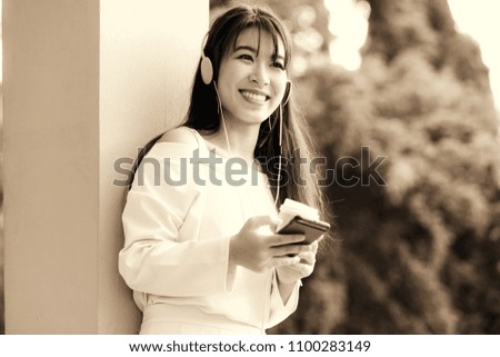 Happy asian girl with headphones listening to music from mobile phone drinking coffee. Relaxed woman holding mobile phone and coffee cup, selective focus color filter effect,copy space.