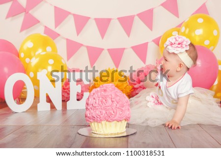 Portrait of adorable Caucasian baby girl in tutu tulle skirt celebrating her first birthday. Cake smash concept. Child kid crawling on floor in studio with pink flags and balloons, eating dessert 