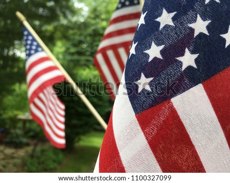 Close up view of a red white and blue American flag with stars and stripes. Two other flags faded into the background with green trees.