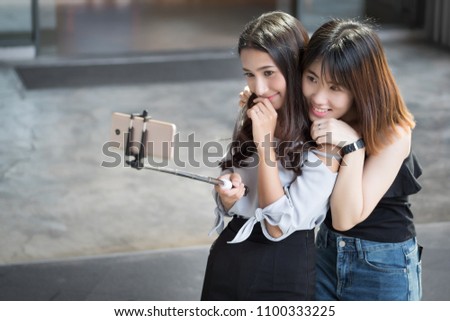 happy woman best friend taking selfie photograph together; smiling asian girl with her bestie using selfie stick to take hobby, leisure, casual selfie photo in urban environment; asian 20s woman model