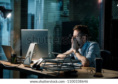 Man covering mouth while yawning at table in dark office having overtime.  Royalty-Free Stock Photo #1100338646