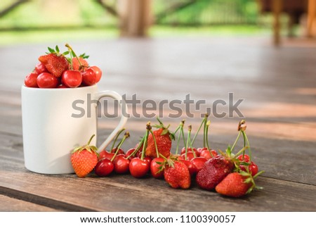 A white cup with fresh berries: cherries, strawberries, blueberries and raspberries, with more blurred berries in the background , shot from above on a light wooden background texture