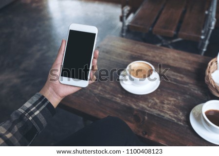 Mockup image of a woman's hand holding white mobile phone with blank desktop screen with coffee cups on wooden table in cafe