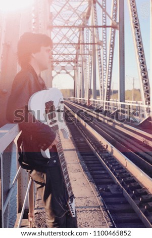 A man with an electric guitar on the railway. A musician in a leather jacket with a guitar on the street in the industrial zone. Guitarist on the bridge.
