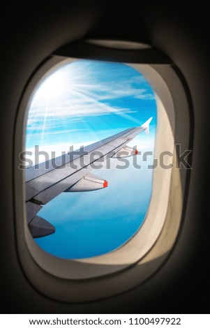Wing of an airplane photographed through the porthole window, with blue sky, clouds and sun rays
