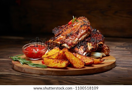 Fried ribs with rosemary, potatoes rustic, onion, sauce on wooden round Board. Dark background. Place for text, copyspace Royalty-Free Stock Photo #1100518109