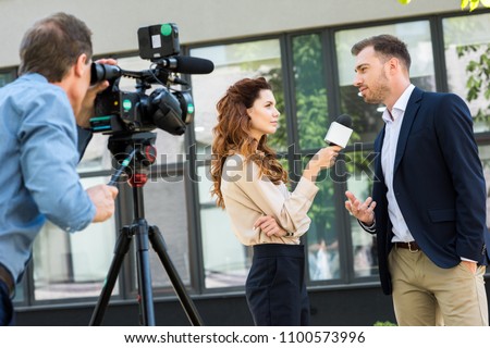 professional cameraman and journalist interviewing businessman near office building  Royalty-Free Stock Photo #1100573996