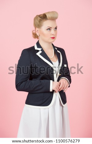 Serious blonde girl in jacket looks up. Woman in pin-up style dreaming standing in studio with closed hands. Blond female with styled make-up