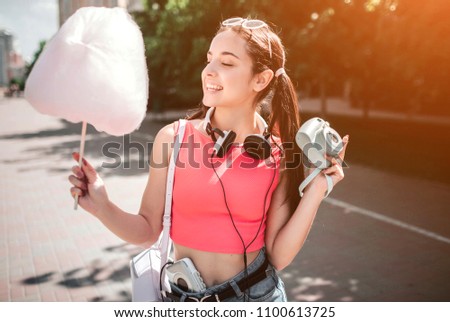 Nice girl is posing. She is holding cotton candy in one hand and camera in the other one. Also there is headphones around her neck and music player in pant. She looks full of joy.