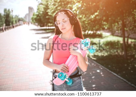 Good-looking girl is standing on street and holding skate in her hands. It is coloured by pink and blue colors. Girl is holding it with both hands and looking down to the side. She is enjoying music.