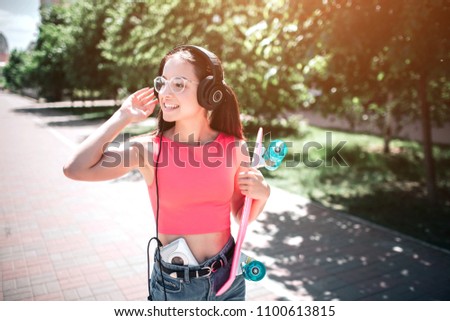 Old-fashioned girl is standing outdoors and listening to music through headphones. She is holding her right hand close to one og headphones and looking to the side. Also she has a skate in left hand.