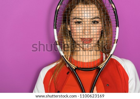beautiful asian girl looking at camera through tennis racquet isolated on violet