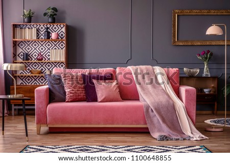 Front view of a pink sofa with pillows and blanket, vintage cupboard in the background in a glamorous living room interior Royalty-Free Stock Photo #1100648855