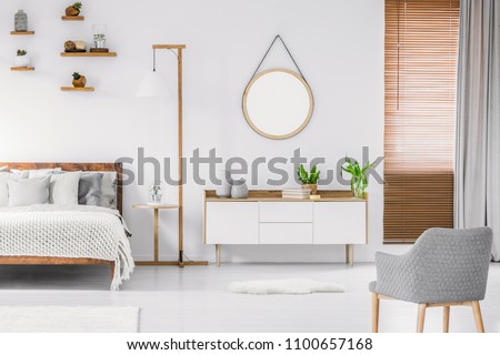 Scandinavian style white room interior with round mirror on the wall, wooden bed with pillows and blanket, cabinet and armchair. Real photo. Royalty-Free Stock Photo #1100657168