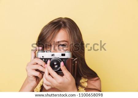 Close up portrait of a pretty young girl taking picture with a photo camera isolated over yellow background