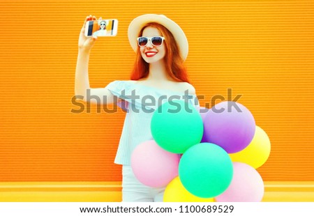 Stylish happy girl an air colorful balloons takes a picture self portrait on a smartphone on orange background