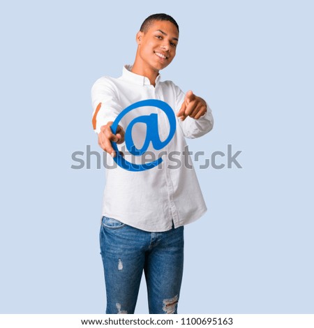 Young african american man with white shirt holding icon of at dot com and pointing to the front on isolated blue background