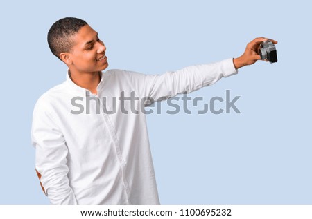 Young african american man with white shirt making selfie with old camera on isolated blue background