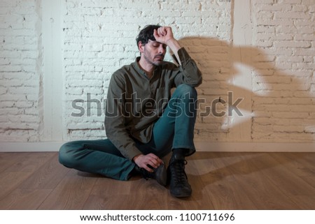young depressed man sitting against a white wall at home with a shadow on the wall feeling miserable, lonely and sad in mental health depression concept Royalty-Free Stock Photo #1100711696