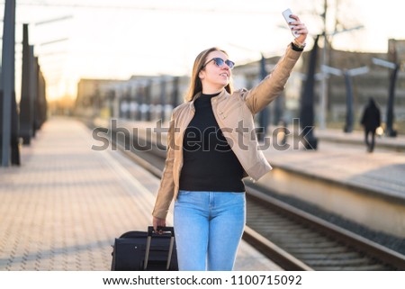 Woman taking selfie with mobile phone in train platform at station. Smiling and happy lady taking photo of herself with smartphone. Traveler and tourist arrived to destination.