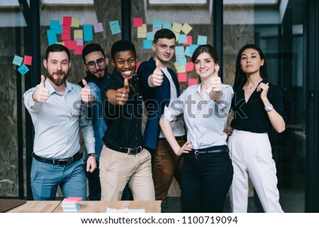 Portrait of multicultural team of happy office employees dressed in formal wear holding thumb up showing sign okay and success of collaboration standing against wall with colorful stickers in office