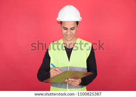 Engineer man wearing a helmet and holding a pen and clipboard in his hand and he is writing something, standing on a red background.