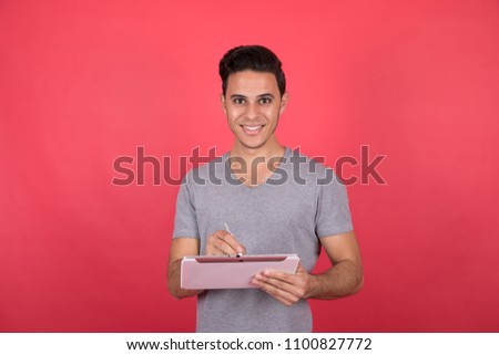 Handsome young man wearing a casual outfit, holding a tablet and the pen writing something and smiling, standing on a red background