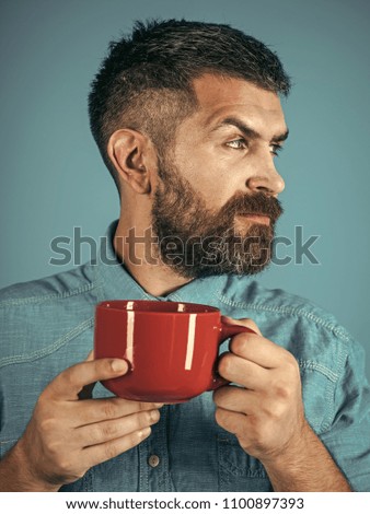 Man with serious emotion. Bearded hipster with milk cup. Refreshment and energy. Red mug with cocoa, hot wine or chocolate. Guy with mulled wine on blue background. Man drink morning coffee or tea.