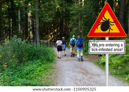 Warning sign "beware of ticks" in infested area in the green woods with hikers Royalty-Free Stock Photo #1100905325
