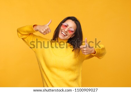 Young shining model in yellow sweater and sunglasses showing thumbs up looking at camera.  Royalty-Free Stock Photo #1100909027