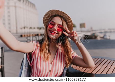 Charming girl in pink sunglasses having fun in city and making selfie. Wonderful blonde young woman taking picture of herself on town background.