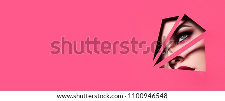 the face of a young beautiful girl with a bright make-up and with plump red lips peeks into a hole in pink paper,Fashion, beauty, make-up, cosmetics, beauty salon, style, personal care, geometry.