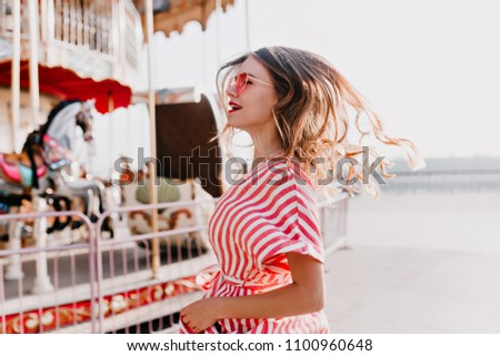 Inspired young woman dancing near carousel. Cheerful caucasian girl in striped dress expressing happiness in amusement park.