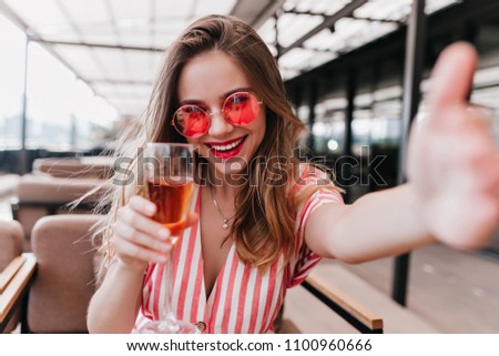 Cheerful trendy girl making selfie while drinking cocktail. Indoor photo of positive female model in pink sunglasses having fun in cafe.